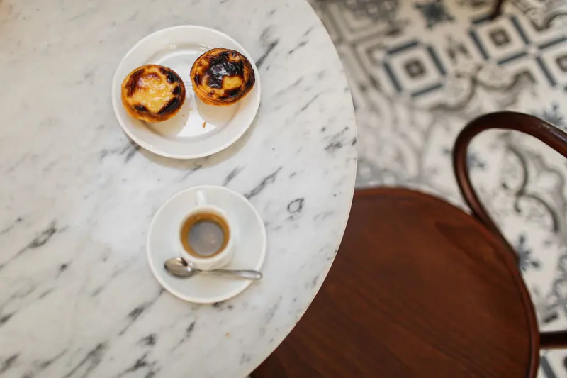 Seen from above, an espresso cup and a plate with two pastéis de nata sit on a round marble table beside a bentwood chair and a patterned tile floor.